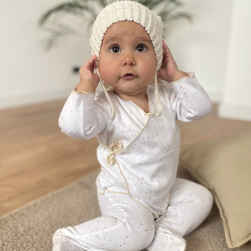 A baby sitting on the floor wearing a white organic wrap top with long sleeves and a V-neck, paired with footed pants that have an elastic waistband and a herringbone bow detail.